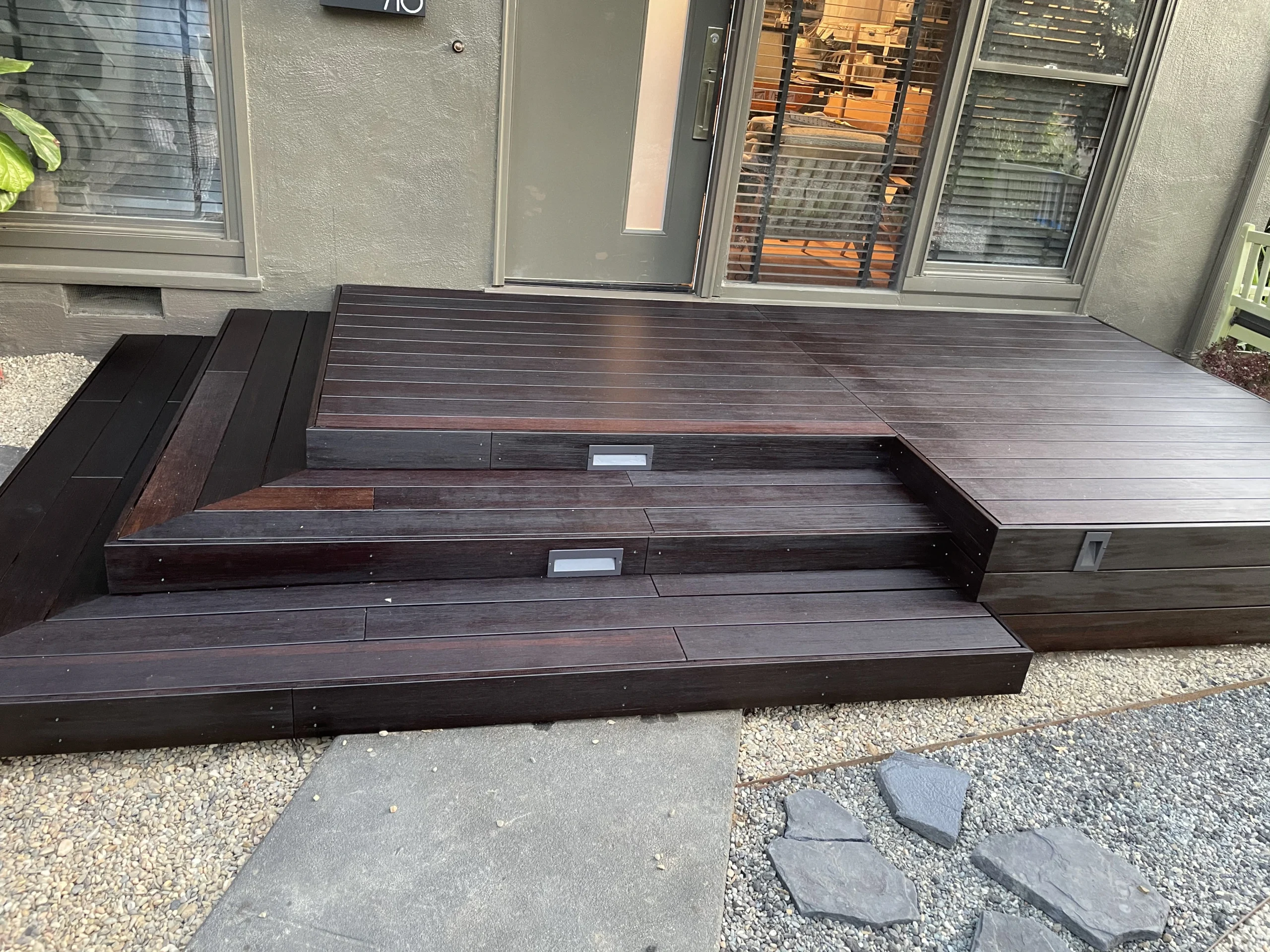 A dark wooden deck with steps leading to a glass door. The deck is adjacent to a gray building with windows. There are pathway stones and gravel leading up to the deck.