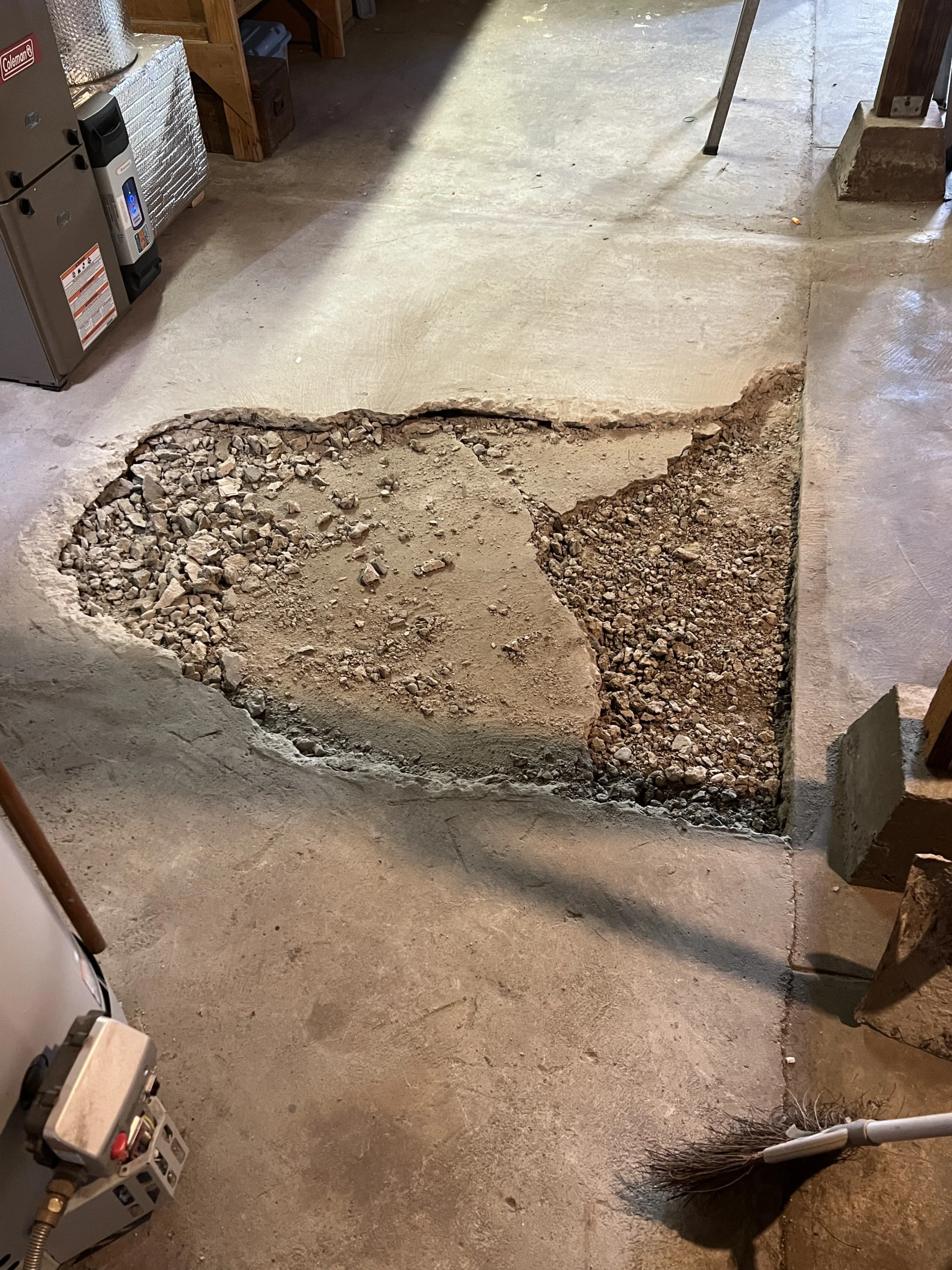 Concrete floor with a partially removed section revealing gravel and broken pieces beneath. A broom is placed nearby on the right. Various tools and equipment are visible in the background.