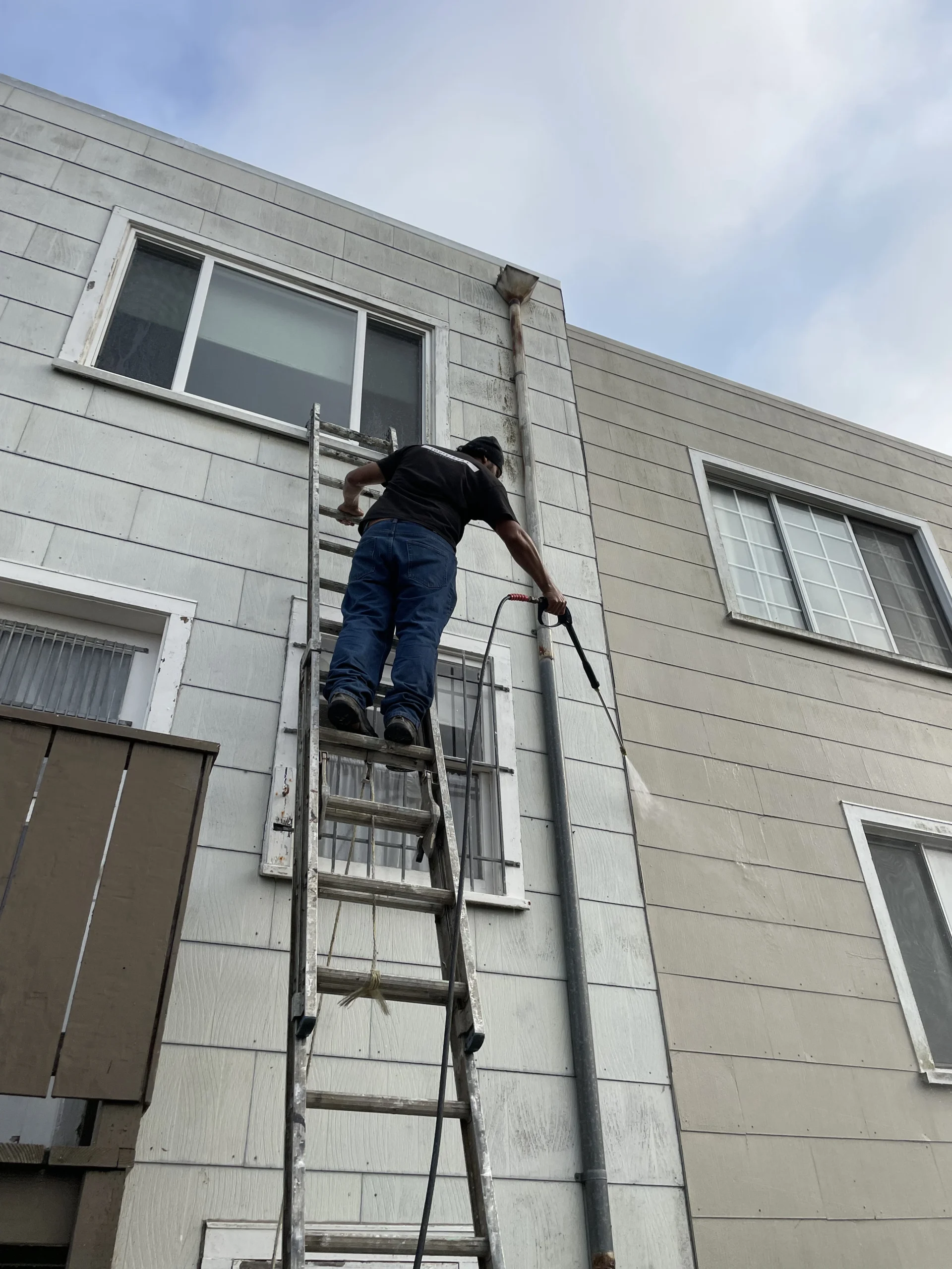 A man climbs a tall ladder against a gray building while holding a hose.
