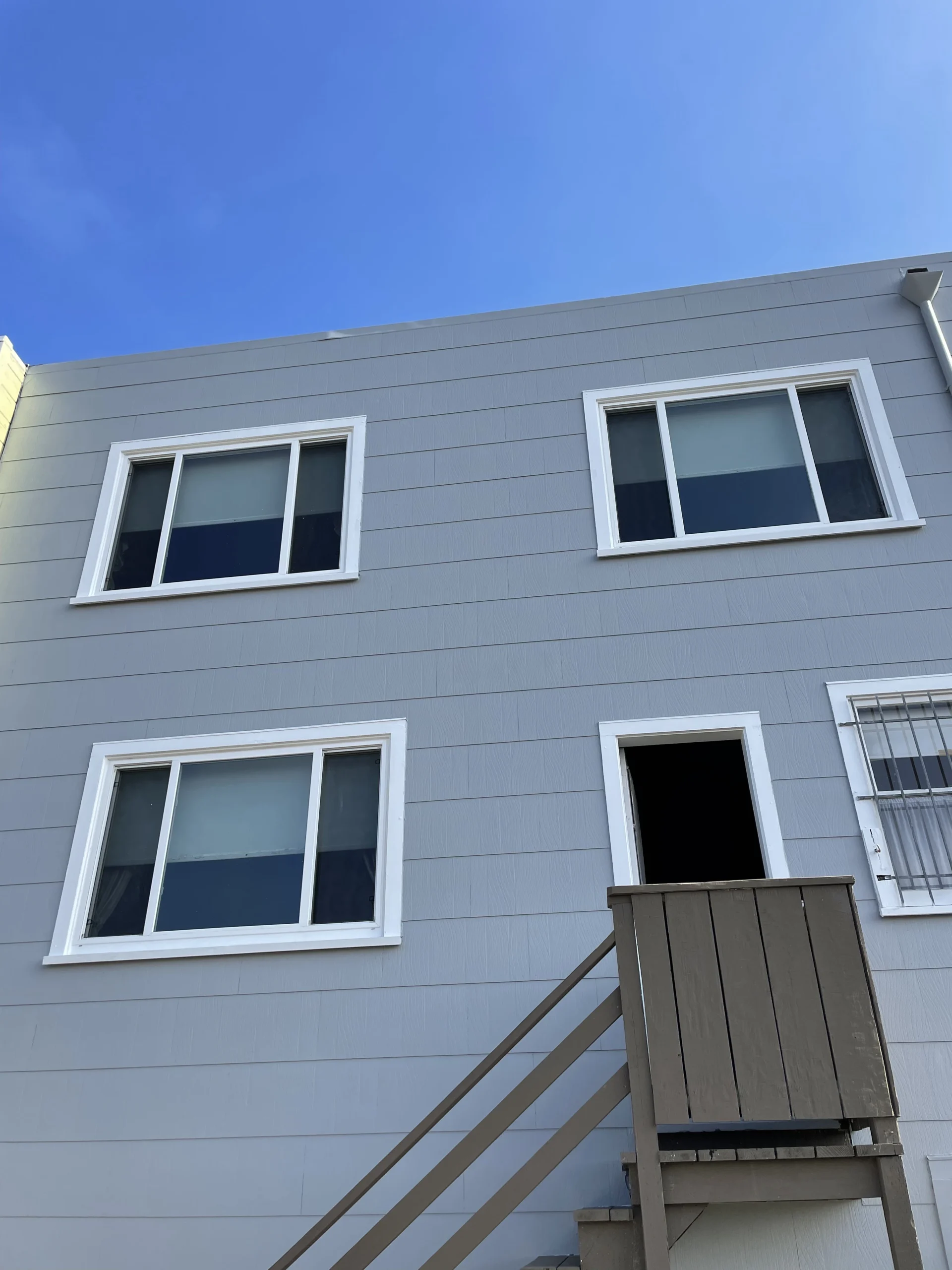 Exterior of a modern two-story building with a wooden staircase leading to an upper balcony and four windows against a clear blue sky.