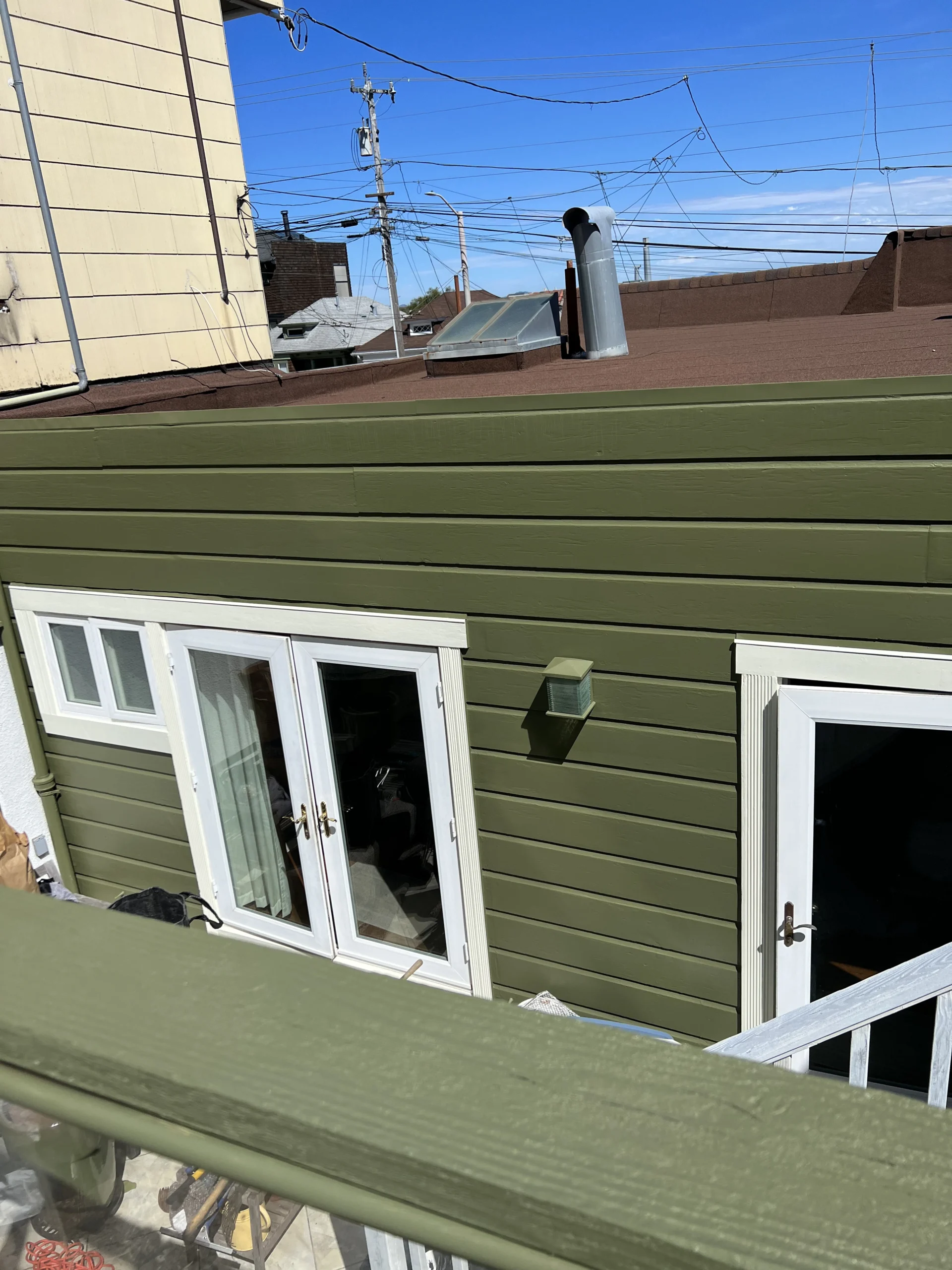 Green siding on a building with white windows, viewed from an upper deck under a clear blue sky, with visible electrical wires and other buildings in the background.