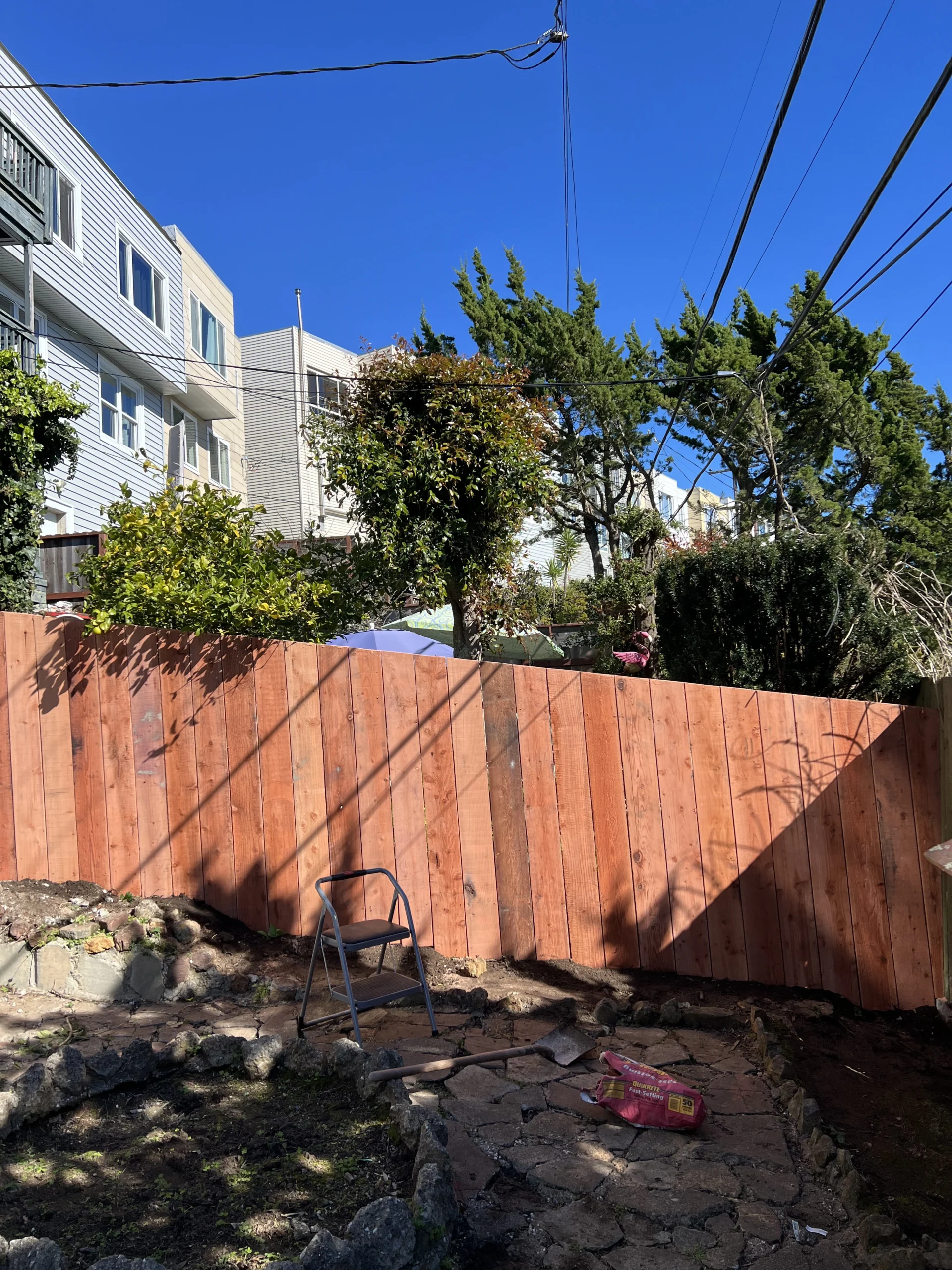 A backyard with a new wooden fence under a clear blue sky. There is a small ladder and a bag of materials next to the fence. Trees and neighboring buildings are visible in the background.
