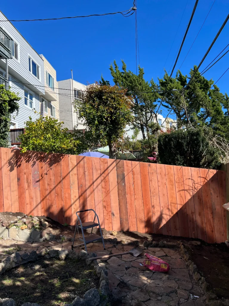 A backyard with uneven wooden fencing, a metal step stool, and a partially paved ground. Residential buildings and trees are visible in the background under a clear blue sky.