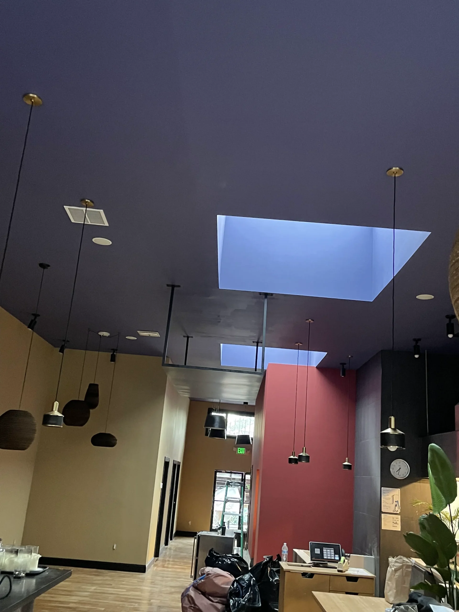 Interior of a modern cafe with a purple ceiling, square and round hanging lights, and red and gray walls. a partially visible counter and bags are in the foreground.