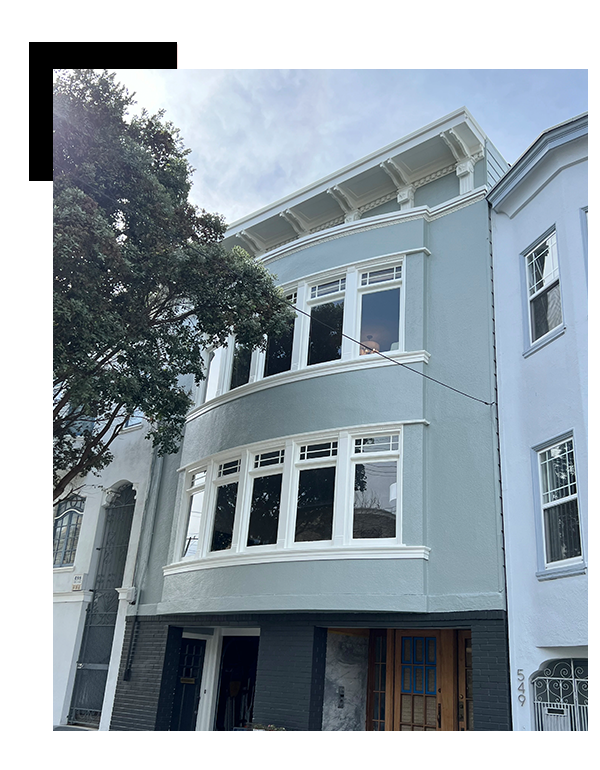 A three-story gray residential building with white trim, located next to a similarly structured blue building. The building features large bay windows and a tree obscuring part of its facade.