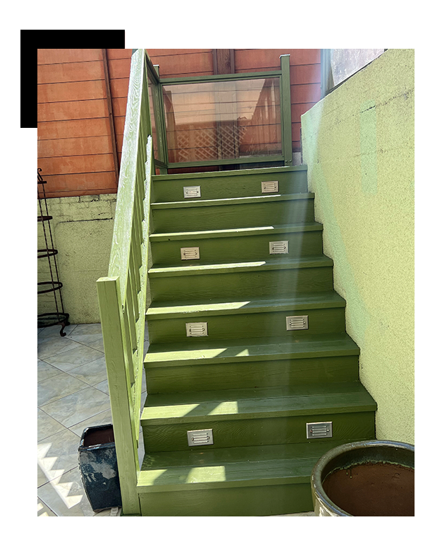 A green-painted wooden staircase with metal step plates, handrails, and a transparent guard at the top. The staircase is located outside, against walls with a mix of green and red-brown colors general carpentry.