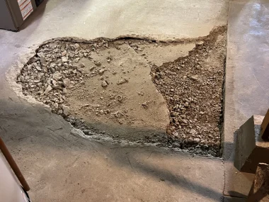 A damaged concrete floor in a garage showing exposed rubble and a jagged hole, with a broom and equipment in the background.