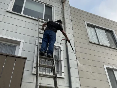 A man climbs a tall ladder against a gray building while holding a hose.