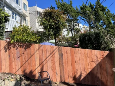 A wooden fence in the foreground of a residential area with visible houses, trees, and power lines under a clear blue sky.